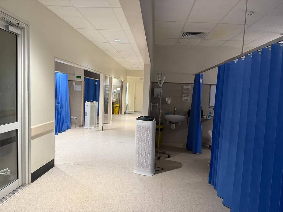 Hospital hallway with blue curtains, empty patient bays, and medical equipment. Bright, clean setting with a calm, sterile atmosphere.