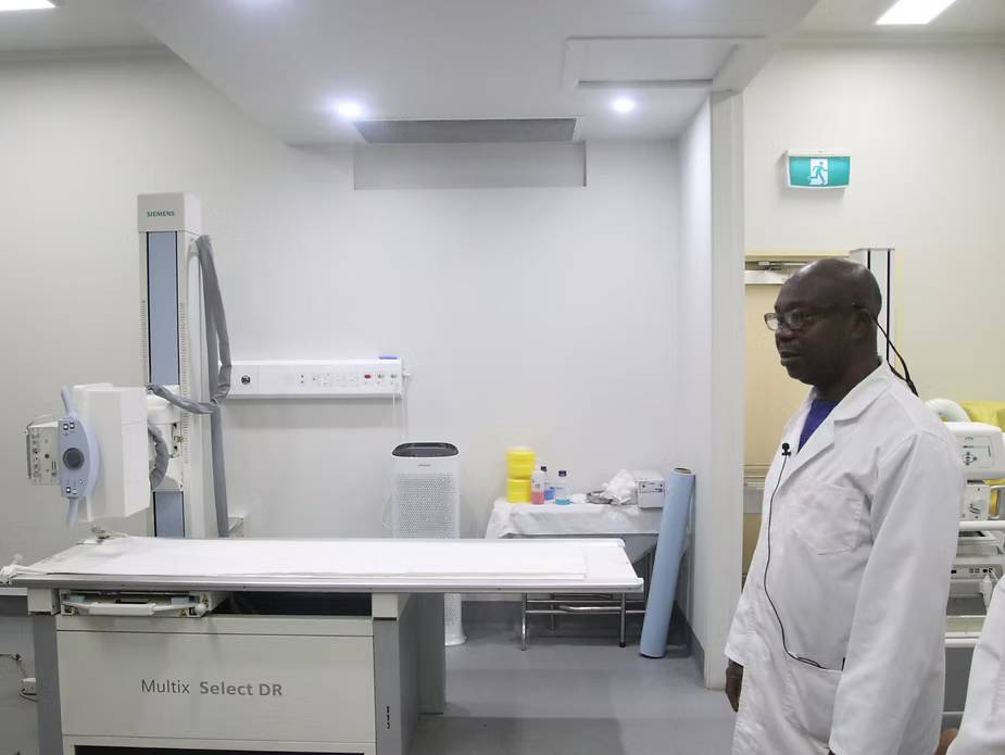 Man in lab coat stands in a white medical room with Siemens Multix Select DR X-ray machine. Bright, clinical setting. Exit sign visible.