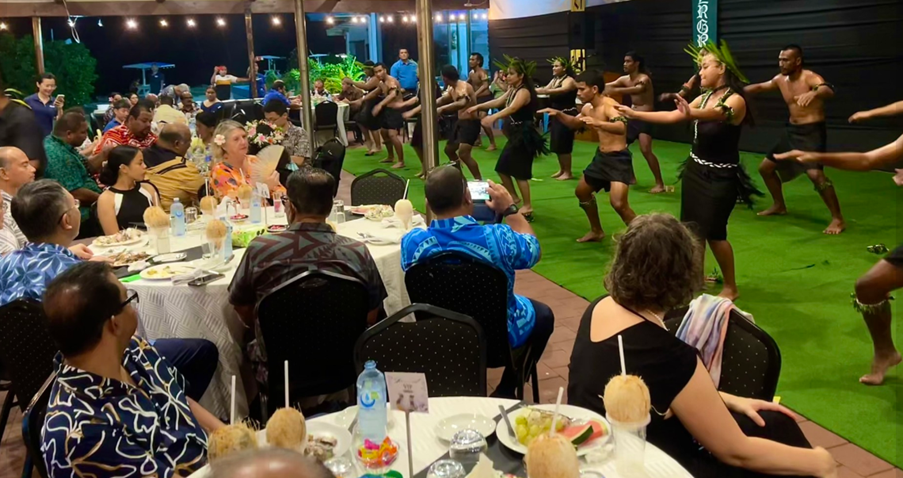 The image depicts a vibrant, outdoor gathering with people dressed in colorful attire, enjoying a meal and socializing, likely in a tropical or festive setting.
AI-generated content may be incorrect.